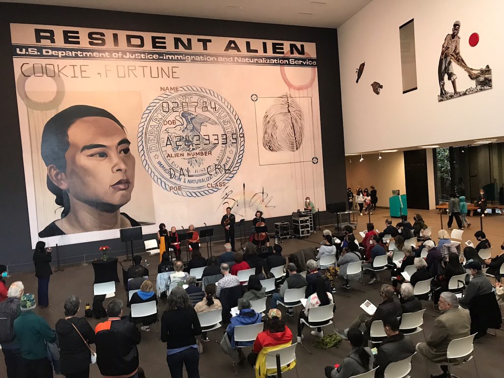 Poet Genny Lim stands before Hung Liu’s mural, Resident Alien, as she reads her poem “My True Name” for the audience seated in the de Young Museum’s Wilsey Court.