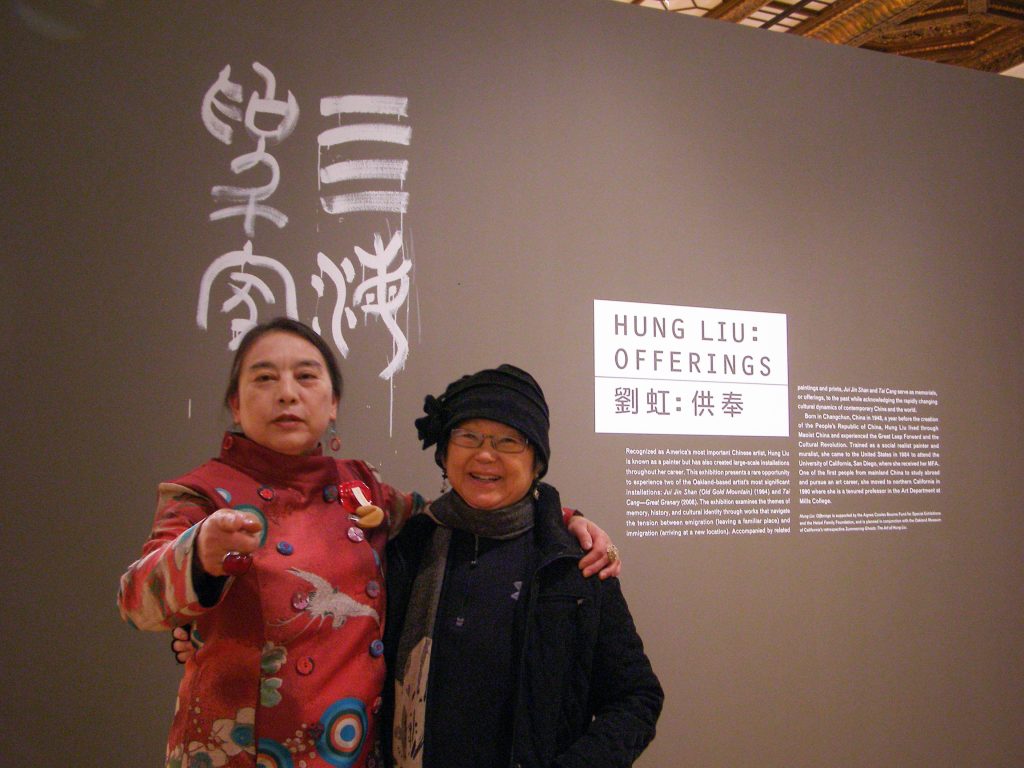 Hung Liu, dressed in red, wraps one arm around artist Flo Oy Wong, as she points towards the photographer (Lenore Chinn). Zuan Shu or Seal Script Chinese calligraphy is painted in white, with Hung Liu’s characteristic drip, on the gray wall with the exhibit text for “Hung Liu: Offerings.”