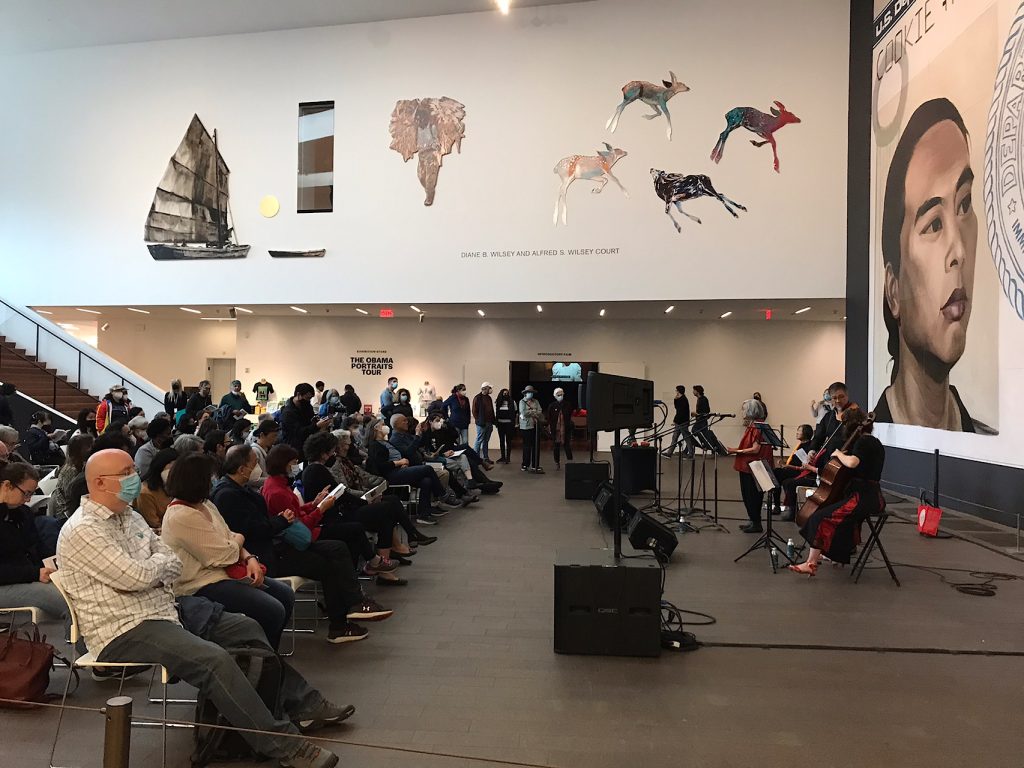 Poet Nellie Wong stands before Hung Liu’s mural, “Resident Alien,” as she reads her poem “Pantoum for the Woman Who Left Cloud Village” for the audience seated in the de Young Museum’s Wilsey Court.