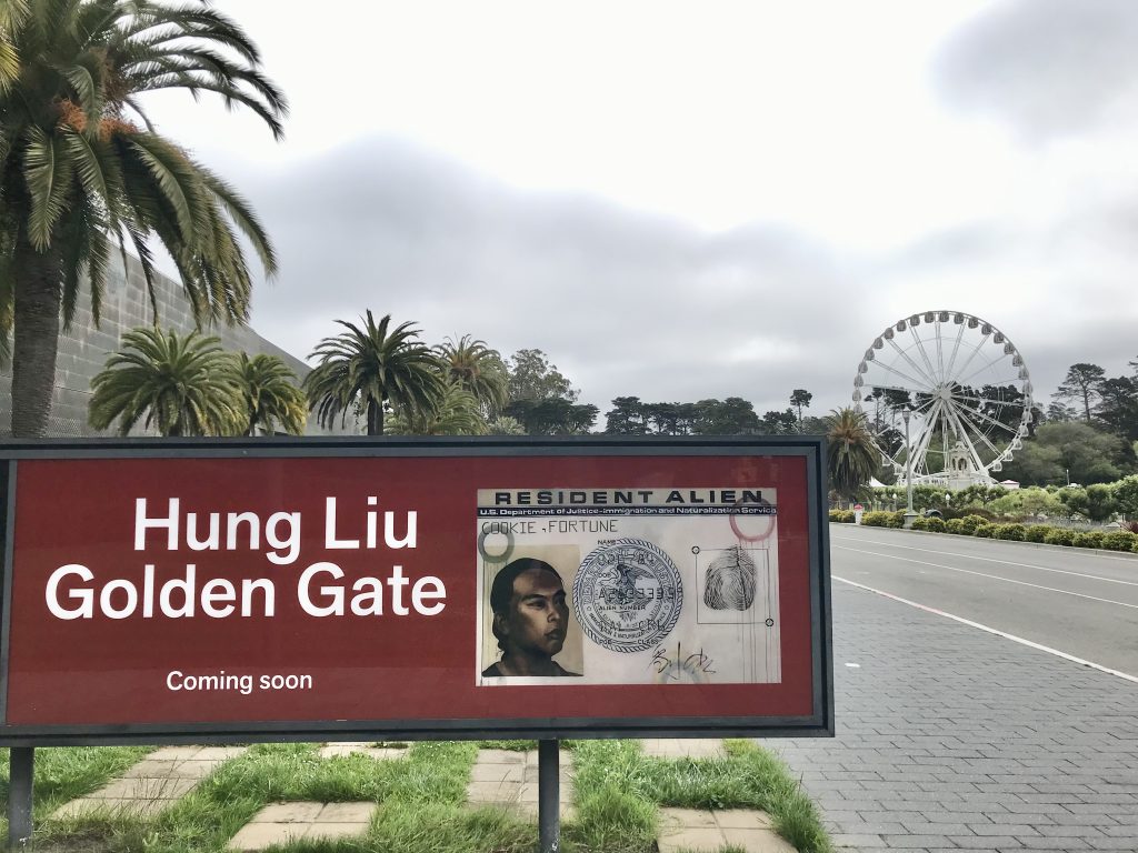 Sign announcing the upcoming exhibit, Hung Liu: Golden Gate at the de Young Museum. In the background, palm trees and the StarSky Wheel line Golden Gate Park's Music Concourse.
