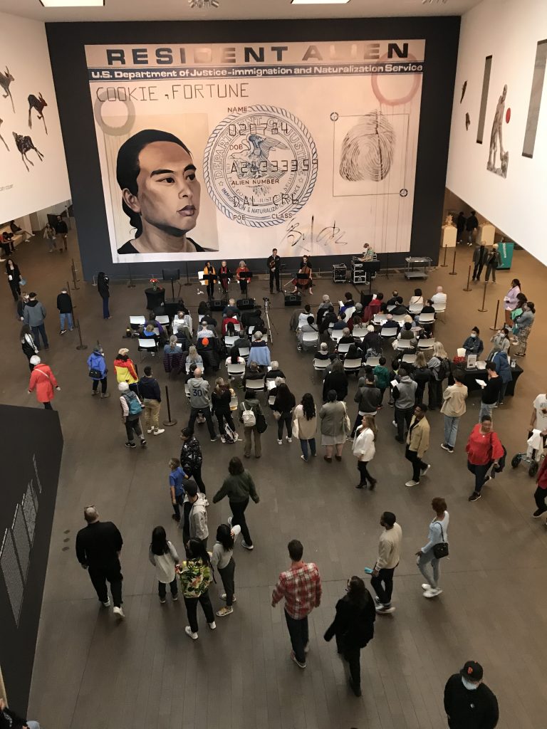 Overhead view of The Last Hoisan Poets poetry performance in the Wilsey Court of the de Young Museum, with Hung Liu’s “Resident Alien” in the background.