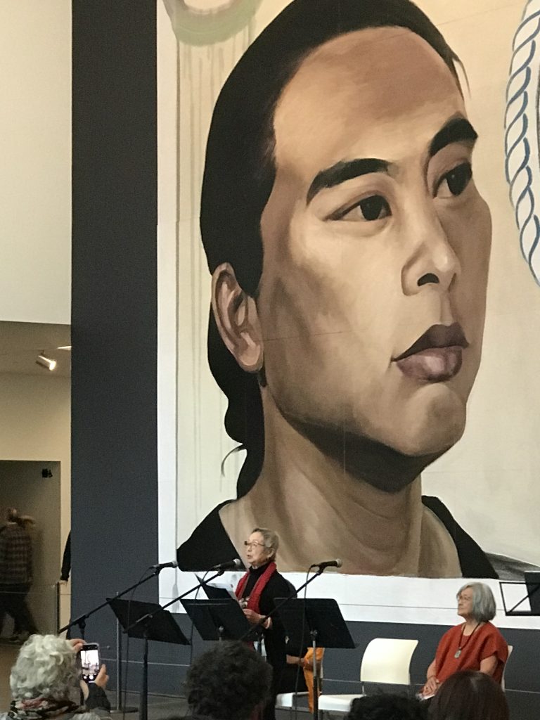 Poet Flo Oy Wong stands before Hung Liu’s mural, Resident Alien, as a seated Nellie Wong looks on. Flo reads her poem “They are the Rays of Her Sun” for the audience seated in the de Young Museum’s Wilsey Court.