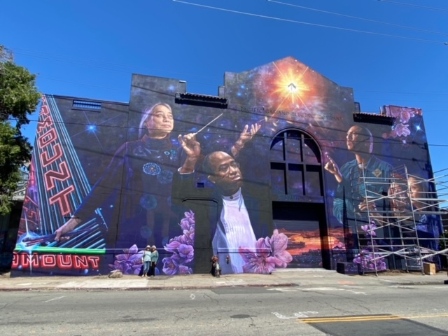 A larger-than-life Hung Liu looks skyward as dandelions float from her dress towards the light, in the mural, “Love Letter to Oakland 2,” which also depicts conductor Michael Morgan and Kev Choice and muralists Elaine Chu and Marina Perez-Wong of the Twin Wall Mural Co. Standing on the sidewalk below the mural painted on the PG&E substation, sisters Flo Oy Wong and Nellie Wong, are dwarfed by gigantic purple blossoms.