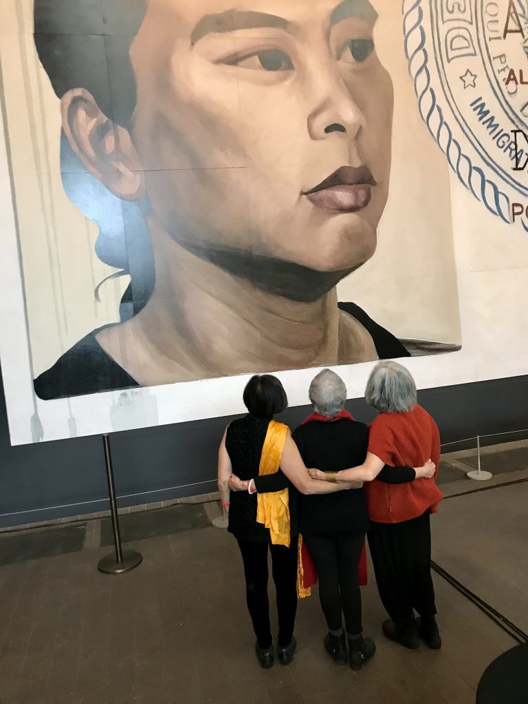 The Last Hoisan Poets - Genny Lim, Flo Oy Wong & Nellie Wong - with backs turned and arms linked, stand facing Hung Liu’s self portrait in the large-scale mural, “Resident Alien,” installed in the de Young Museum’s Wilsey Court.